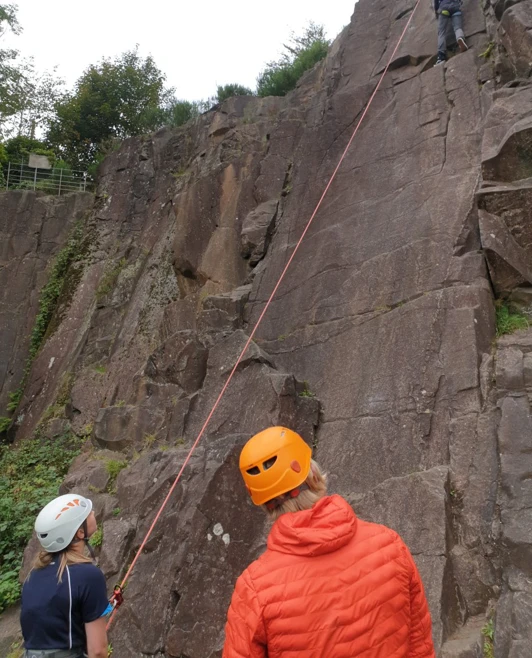 Two students supervise a third climbing a tall rock wall with a safety rope during an outdoor adventure activity. Two students supervise a third climbing a tall rock wall with a safety rope during an outdoor adventure activity.